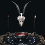 A goat skull with horns above a bowl of red liquid, flanked by two black candles on a dark cloth with mystical symbols.