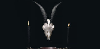 A goat skull with horns above a bowl of red liquid, flanked by two black candles on a dark cloth with mystical symbols.