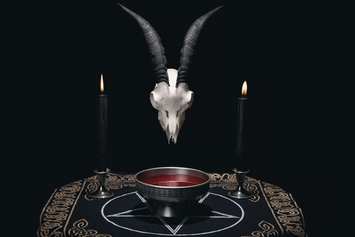 A goat skull with horns above a bowl of red liquid, flanked by two black candles on a dark cloth with mystical symbols.