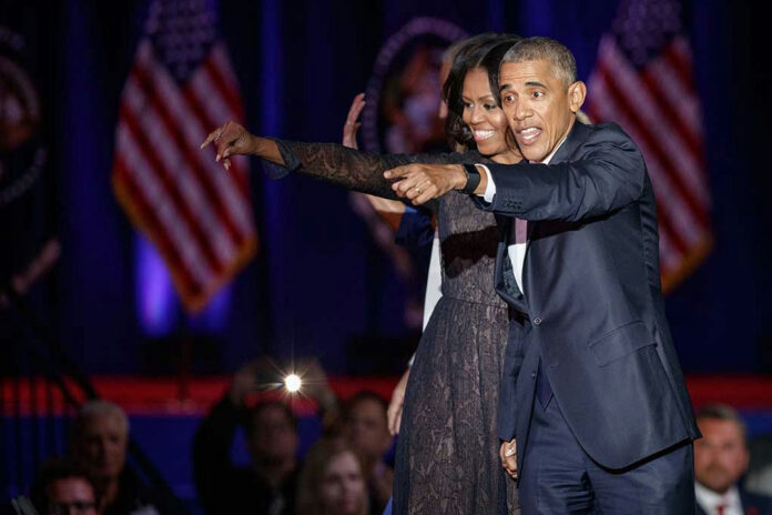 Two people pointing and smiling on stage, American flags behind.