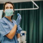 Nurse wearing scrubs and mask putting on gloves.