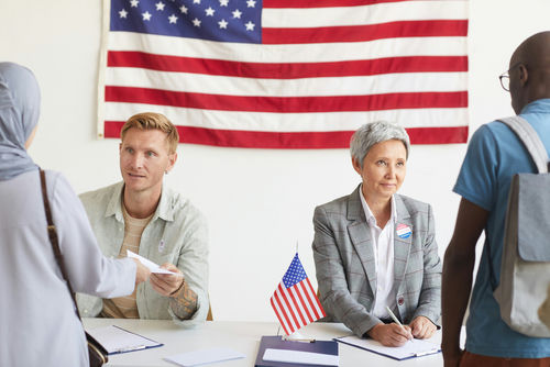 Election workers assist voters at a registration table with a U.S. flag behind them