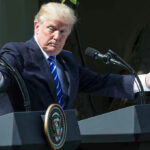 Final Call Haunts Trump After Lindsey Graham’s Shock Death Man speaking at a podium, arms extended outward.