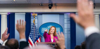 Press secretary at podium, answering reporters questions, raised hands.
