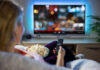Trump Broadcast Blackout, Licenses Threatened Person watching TV with remote, holding popcorn.