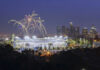 Fireworks above a brightly lit stadium at night with city skyline