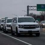 A convoy of white SUVs on a highway with a restricted access sign