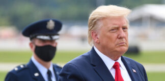 Donald Trump in suit with military personnel behind him.